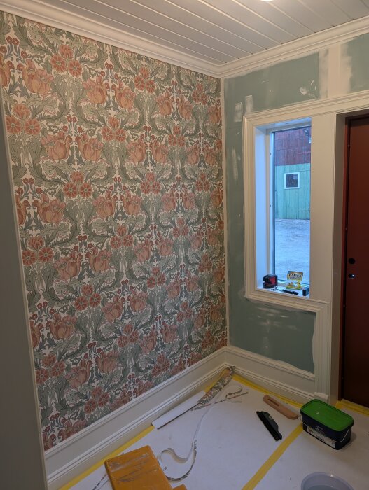 Vägg med mönstrad tapet i rum under renovering, verktyg och färgburk på golvet, del av väggen vid fönstret saknar tapet.