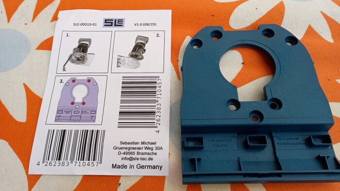 Blå plastkomponent med hål bredvid ett papper med installationsinstruktioner och streckkod, placerad på en mönstrad orange duk.