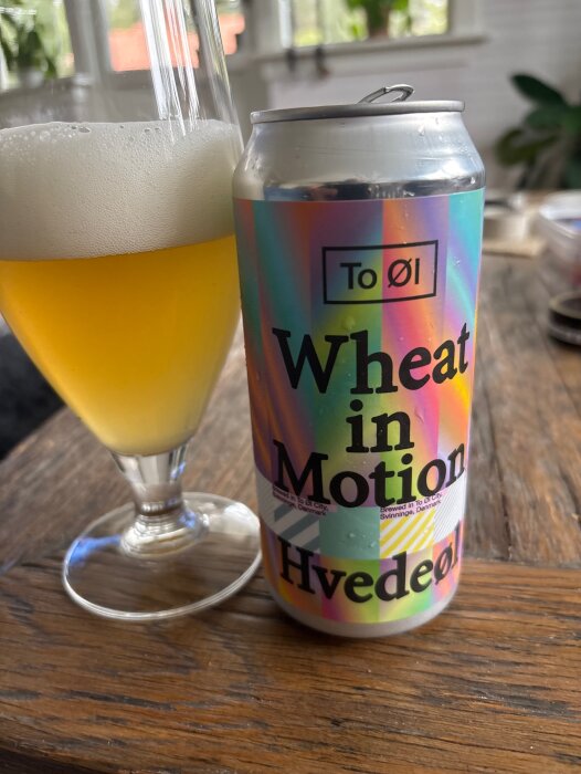 En burk med regnbågsfärgad etikett som visar "Wheat in Motion" bredvid ett glas ljus veteöl på ett träbord.