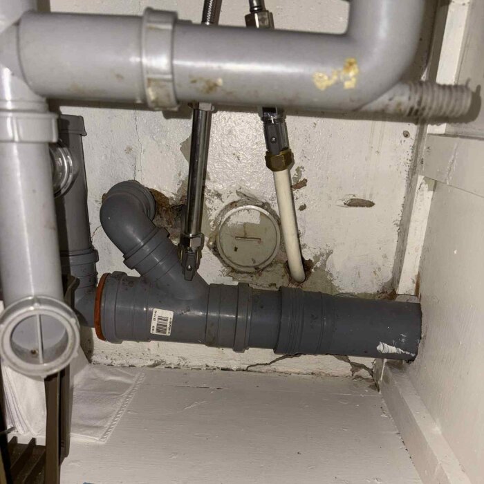 Grå avloppsrör och ventiler i ett trångt utrymme, med synligt slitage på väggen bakom, kopplade till fastighetens ventilation.