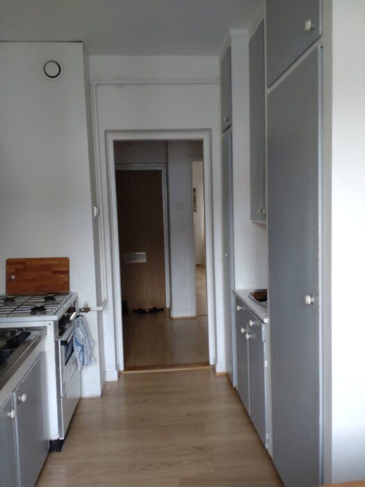 Kök med gråa skåp, spis, träskiva och upphängd kökshandduk, sett från matplats mot hall och sovrum.