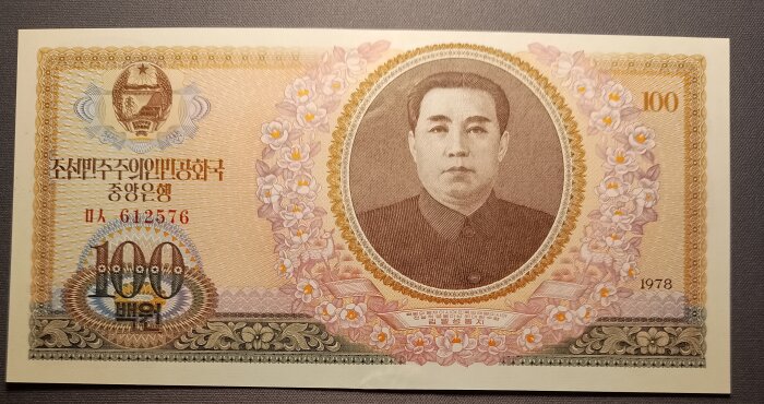 100 won sedel med porträtt av en ledare, omgiven av dekorativa blommor och text på koreanska, utgiven 1978.