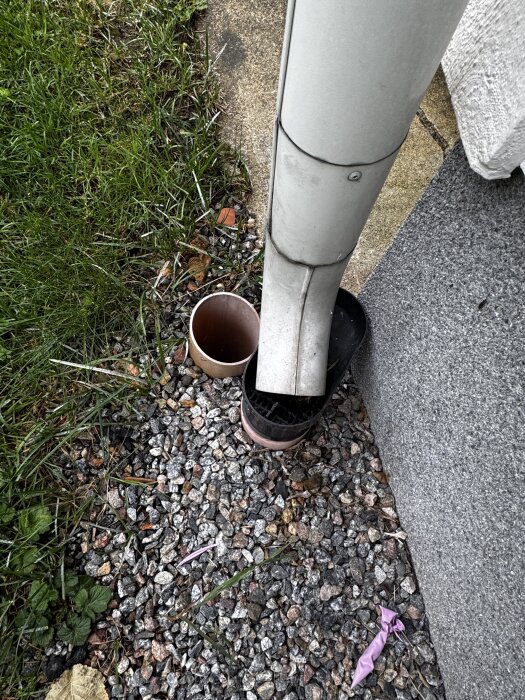 Stuprörsändan med rund öppning intill husvägg, omgiven av grus och gräs, där vattnet leds ner i underjordisk avrinning.