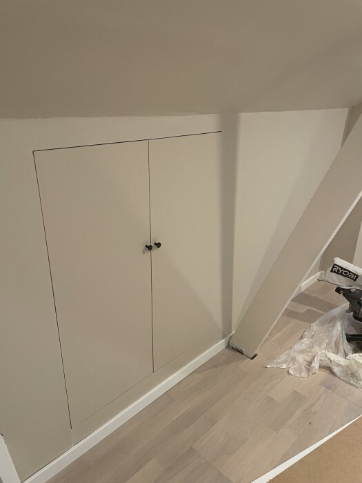 Inbyggda garderobsdörrar under ett snedtak, med ljus trägolv och verktyg synliga i bakgrunden.