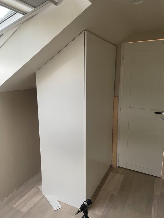 Infälld snedkapad garderob i ett rum med snedtak, målad i vitt. En borrmaskin står på golvet bredvid garderoben.