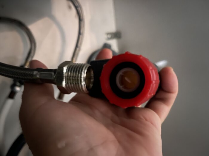 Närbild av en hand som håller en röd aquastop-anslutning till en diskmaskin, med metallgängor synliga.