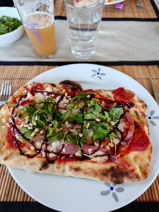 Hemlagad pinsa med tomatsås, skinka, ruccola och pinjenötter på tallrik, omgiven av ett glas öl och vatten.