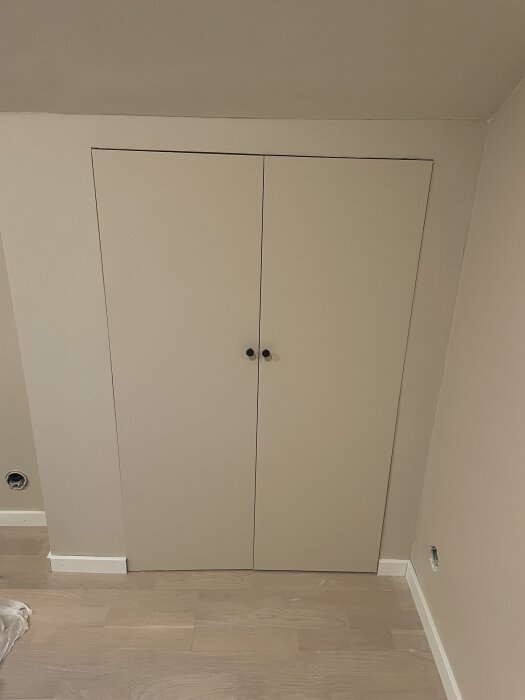Dörrar till inbyggd garderob under snedtak, med ljusbeige yta och två svarta knoppar, monterad mot en beige vägg och trägolv.