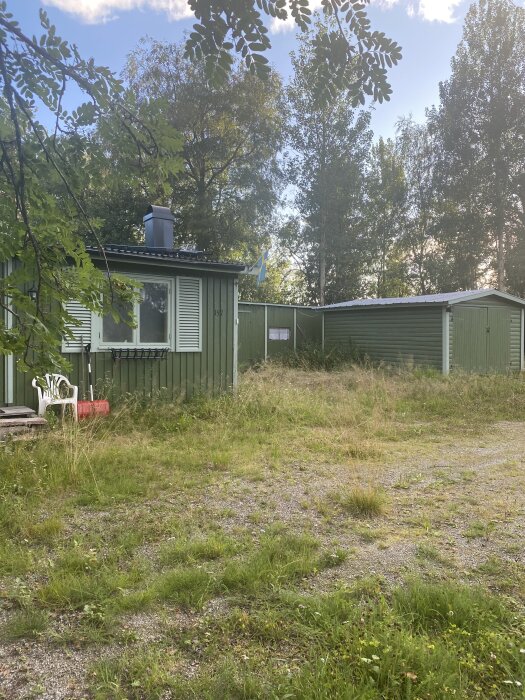 Liten grön stuga med flagga, omgiven av träd och övervuxen trädgård, under blå himmel.