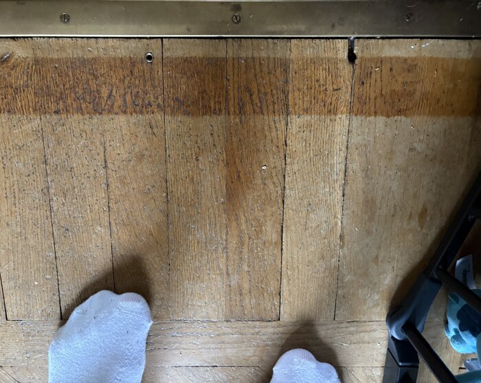 Sliten trägolvsyta med patina, en springa där det verkar ha suttit en gänga, synligt ihop med en sockbeklädd fot och en del av en möbel.
