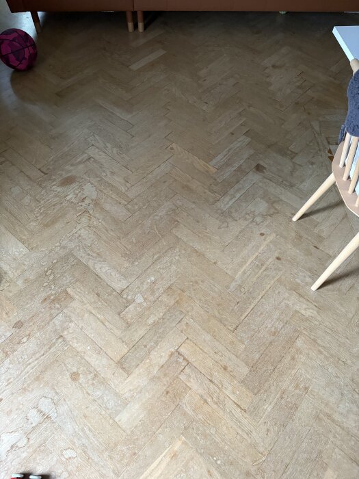 Parkettgolv i fiskbensmönster med synlig patina och en basketball synlig i övre vänstra hörnet. Stolben och bordshörn syns till höger.