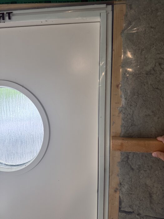 Dörrkarm med glesen under installation, rund fönsterglugg i dörren och isolering synlig på sidan.