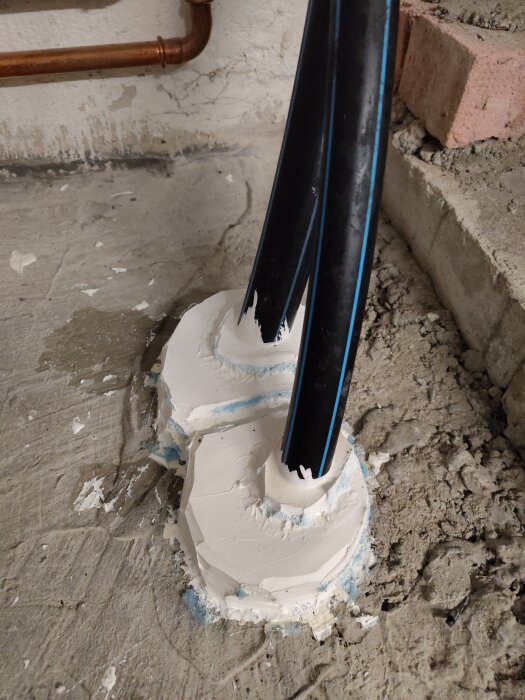 Två svarta rör tätade med vit fogmassa i hård betong, omgivet av röda tegelstenar och en kopparrörsböj, på en betonggolv.