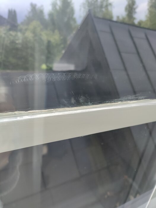 Närbild av ett fönster med cirkulära repor på glaset, ett grått tak och träd i bakgrunden speglas i glaset.