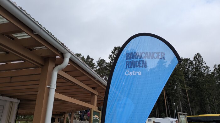Blå flagga med text "Barncancerfonden Östra" framför träkonstruktion och skog.