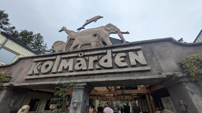 Entré till Kolmården med skulpturer av djur som delfin, elefant, lejon och giraff ovanför skylten. Människor i förgrunden.
