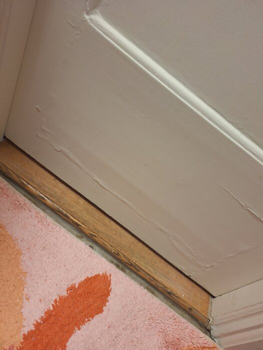 Nederdelen av en vitmålad innerdörr med flagnande färg, trätröskel och rosa-orange matta.