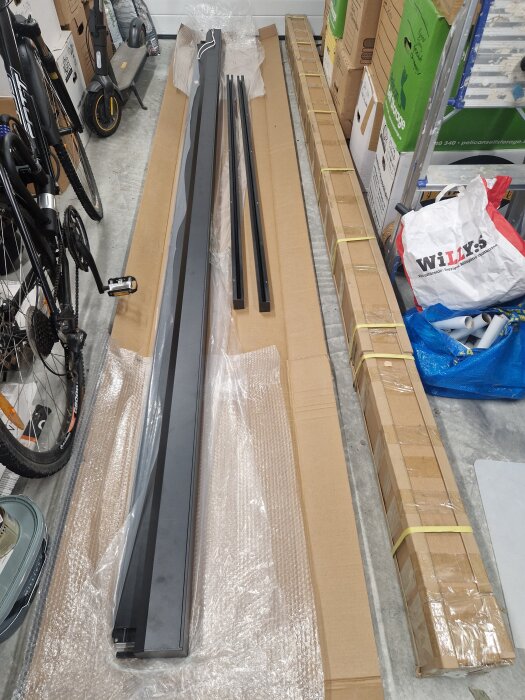 Paket med långa metalldelar och kartong för montering, placerade på golvet nära en cykel och andra paket.