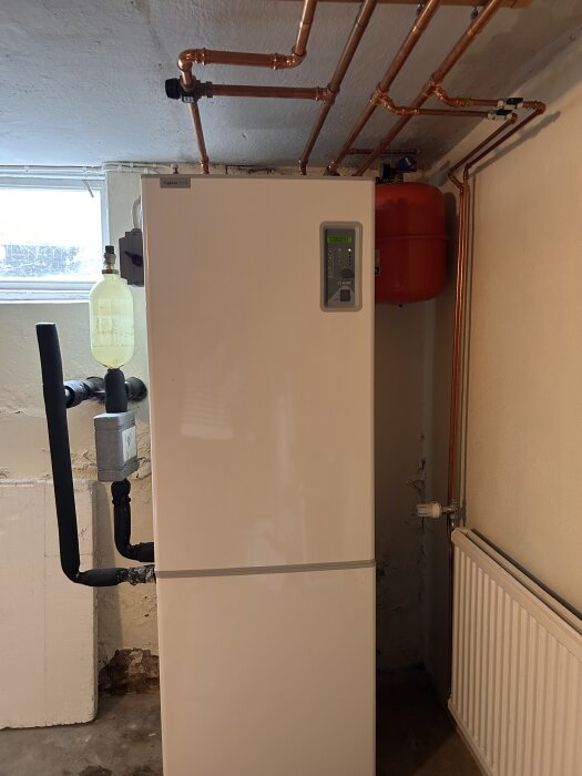 Värmepanna i källaren med etiketten "Fighter 1215", omgiven av kopparledningar och en röd trycktank på väggen bredvid ett fönster.