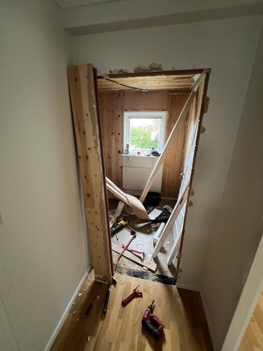 Dörröppning under renovering med exponering av byggelement. Verktyg och brädor spridda på golvet. Fönster som vetter mot trädgård.