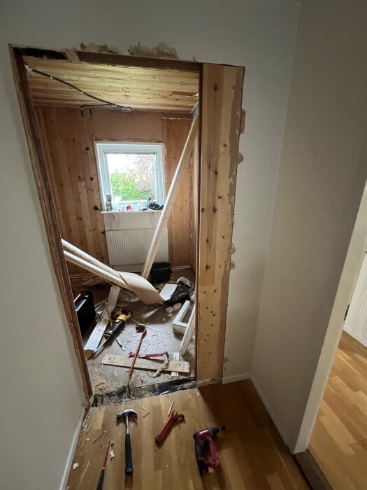 Öppning av ett dörrhål i renoveringsprojekt, verktyg och byggmaterial synliga, träpaneler och ett fönster i bakgrunden.