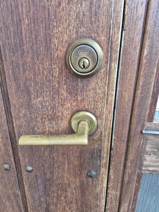 Närbild av en gammal träytterdörr med mässingshandtag och Assa-lås, potentiell installation av Yale Doorman diskuteras.