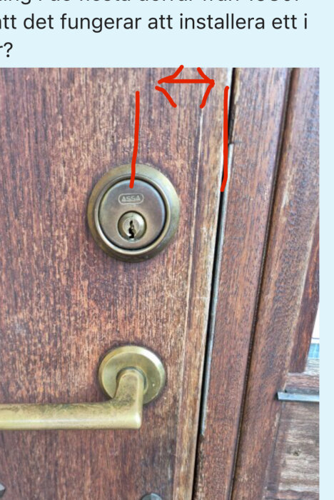 Närbild på en ytterdörr i trä med ett nyckelhål och dörrhandtag; markeringar visar avståndet mellan nyckelhål och dörrkant.