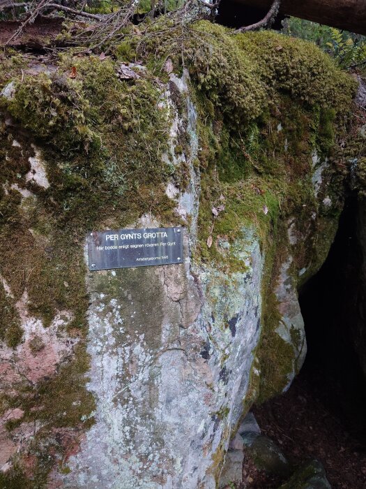 En mossbetäckt klippa med en skylt där det står "Per Gynts Grotta". En mörk öppning syns bredvid skylten.