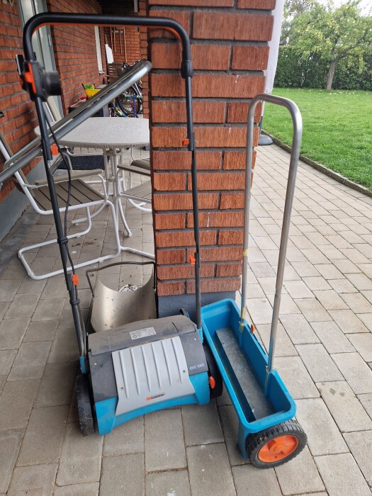 En blå vertikalskärare och gödselspridare står på en stenlagd uteplats mot en tegelvägg.