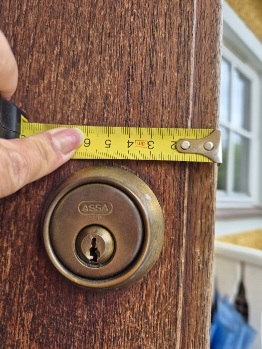 Mäthand som håller ett måttband vid ett dörrlås av märket ASSA på en träyta. Ett fönster syns i bakgrunden.