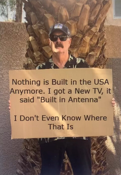 En man i keps och solglasögon håller en skylt med texten om produkter som inte tillverkas i USA längre, enligt skämttexten.