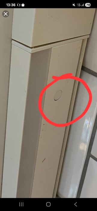 Skruvhål täckt av plastplugg i ett vitmålat elskåp, markerat med en röd cirkel.