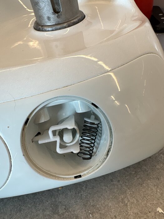 En vit apparatdel med ett synligt fack där en fjäder och en vit plastdetalj finns. Fjädern är löst sittande intill den vita komponenten.