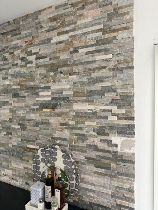 Vägg med stenpanel, delvis borttagen för att avslöja tapet under. Flaska och kryddor på en bänk framför väggen.