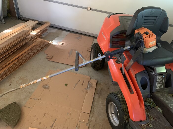 Orange Husqvarna åkgräsklippare i garage bredvid träplankor och kartong.