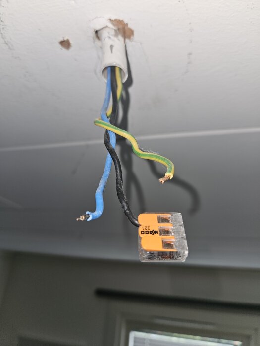 Sladdar hänger från taket med en orange Wago-kopplingsklämma ansluten, visar blå och svarta trådar samt en oisolerad gröngul jordtråd.