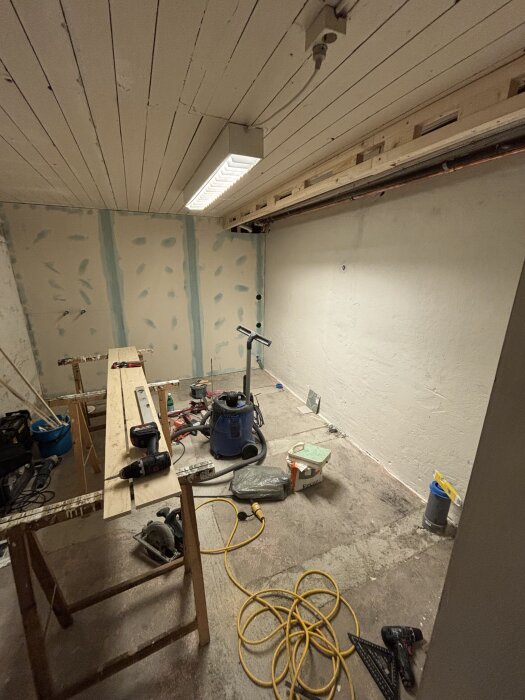 Byggarbetsplats med spacklade gipsskivor, verktyg på bockbord, och byggmaterial vid en vägg där rör låda påbörjats, taklampan lyser.