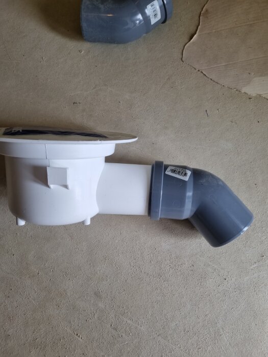 Vit plastbrunn med ansluten grå vinklad rördel ligger på ett cementgolv.