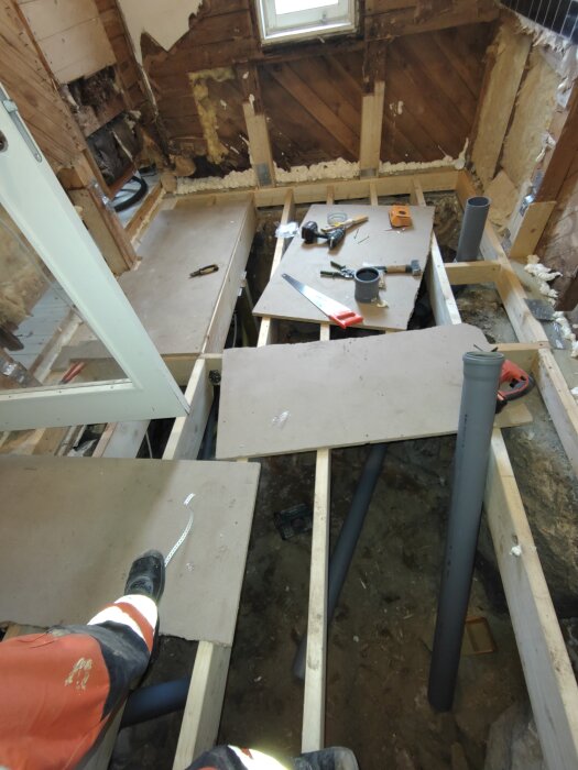 Renovering av badrum med öppet golv, verktyg och byggmaterial synliga, inkl. såg, tejp och rör. Arbetare i orange byxor står på bjälklag.