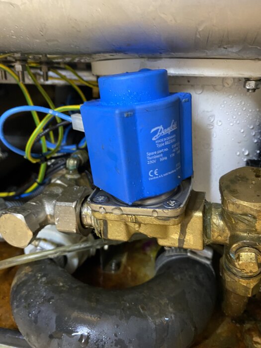 Blå Danfoss ventil under ett rör med vattenläckagefråga, omgiven av gulgröna kablar, mässingsfästen och silverrör.