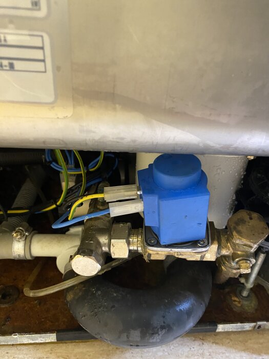 Ventil med blå elektronisk enhet och koppartejp, placerad under en större behållare, omgiven av kablar och rör, eventuellt för reparation.