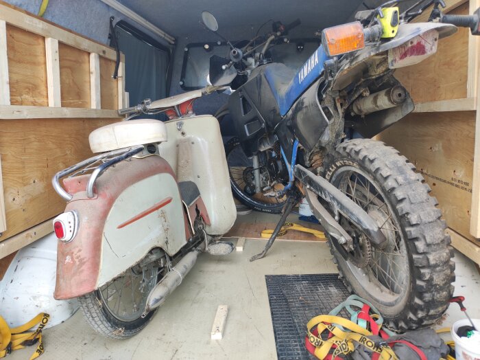 En äldre rödvit moped av årsmodell 1959 och en Yamaha motorcykel står bredvid varandra i ett lastutrymme av trä.