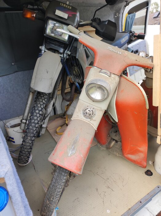 Två mopeder, en rödvit Monark och en grå Yamaha, står parkerade tätt ihop i en bil. Båda är lite smutsiga.