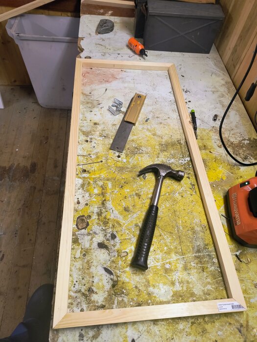 Träram konstruerad av planhyvlade furulister ligger på arbetsbänk med verktyg som hammare och måttstock omkring i en snickeriverkstad.