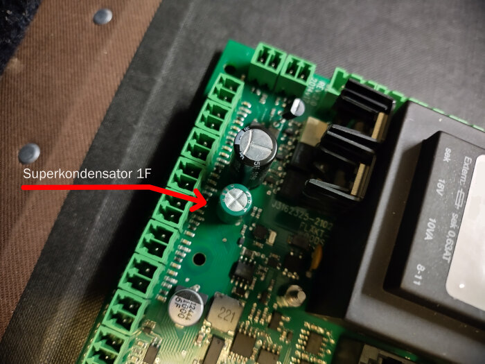 Superkondensator 1F på ett kretskort, markerad med en röd pil och text. Kretskortet har flera anslutningar och komponenter.