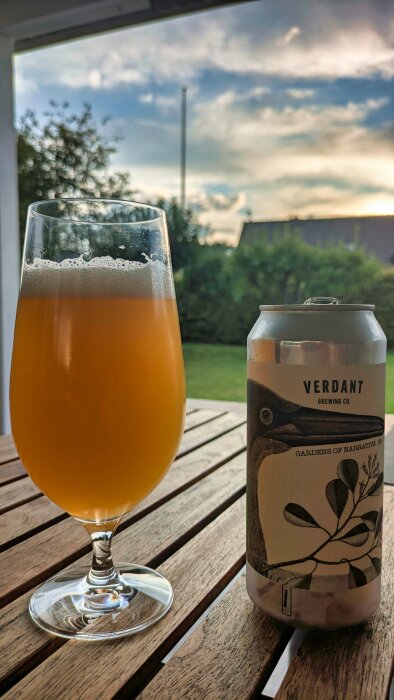 Ölglas med Verdant öl på ett bord utomhus, bredvid en burk med etikett som visar en fågel och blader, solnedgång i bakgrunden.