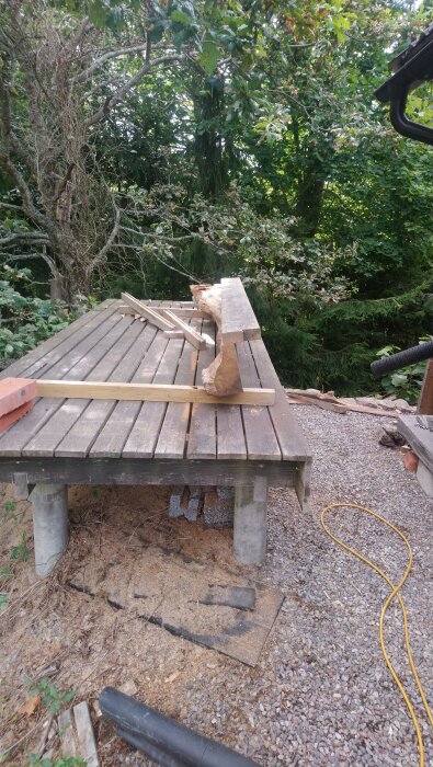 Bitar av ekträ och verktyg på en upphöjd träplattform utomhus, omgiven av träd och grusmark.