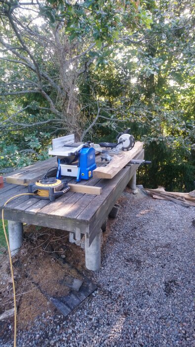 Arbetsbänk utomhus med en såg och en träbit, omgiven av träd och grusmark.
