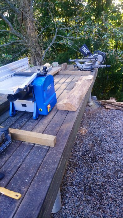 Arbetsbänk utomhus med träbearbetningsmaskiner och en grov ekbit redo för bearbetning, omgiven av träd och grusmark.