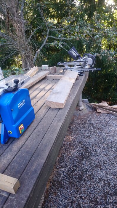 Arbetsyta med såg och träplankor, omgiven av gröna träd och grusmark.